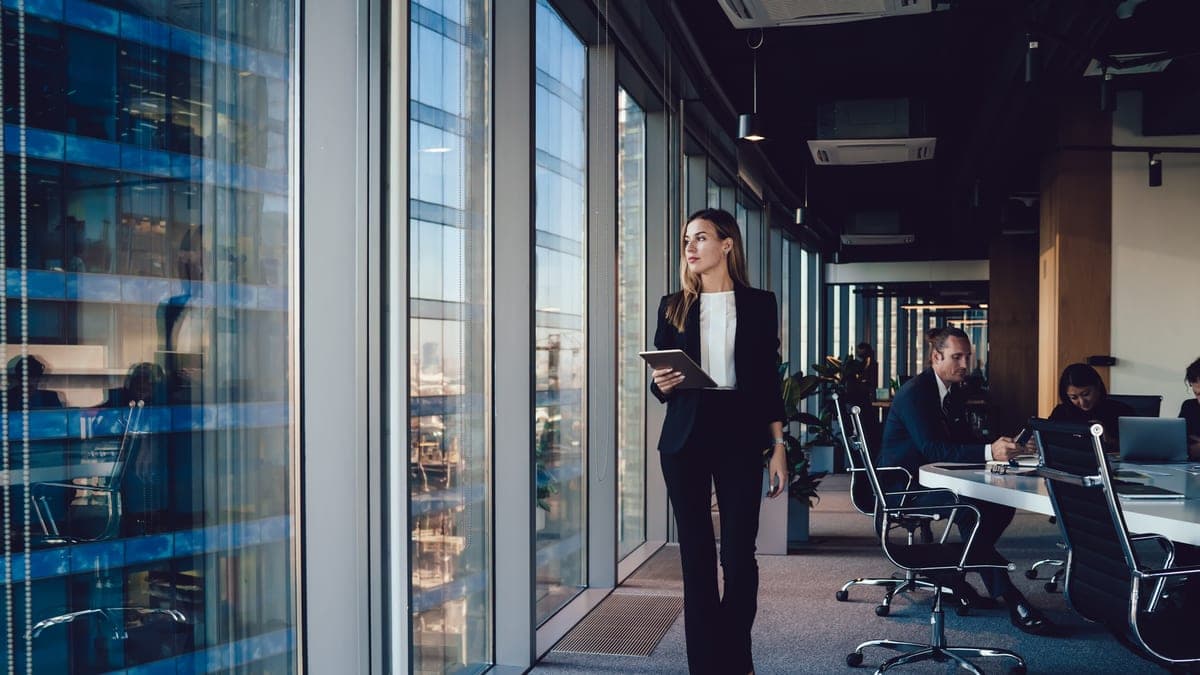 Führungskraft mit Tablet geht durch ein modernes Büro mit Glasfront, Sinnbild für Führung auf Zeit im Interim Management